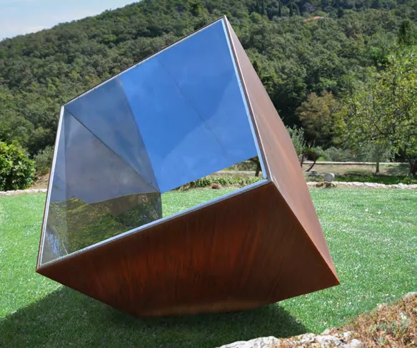 A tilted cube of mirrored stainless steel and Cor-Ten steel standing in a hillside meadow