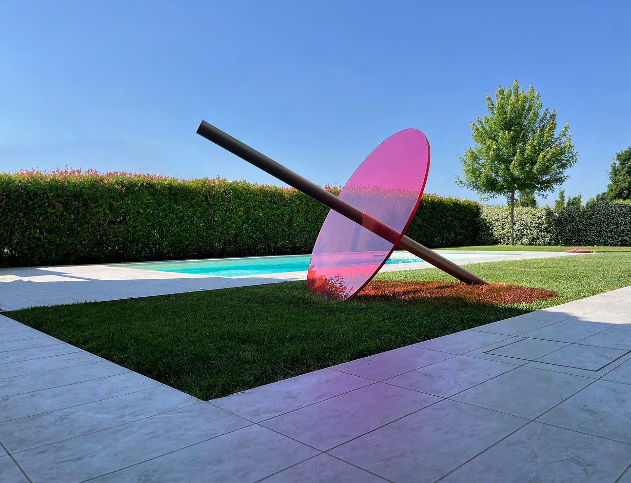 A large translucent magenta glass disc intersected by a dark steel element in a garden
